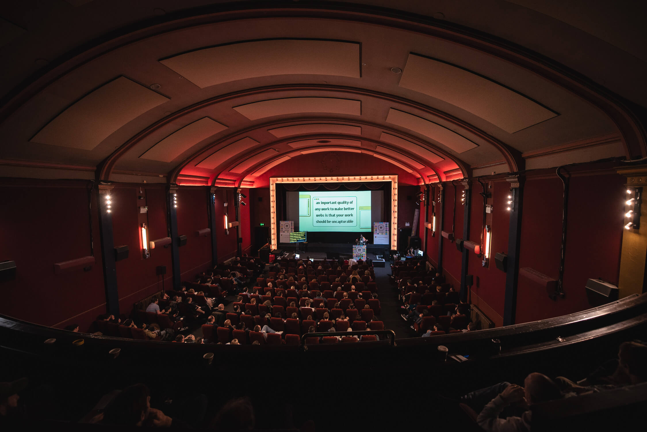 A wide angled photograph from the back of the cinema in the raised seating area, looking towards the stage, where Olu Niyi-Awosusi is opening the conference