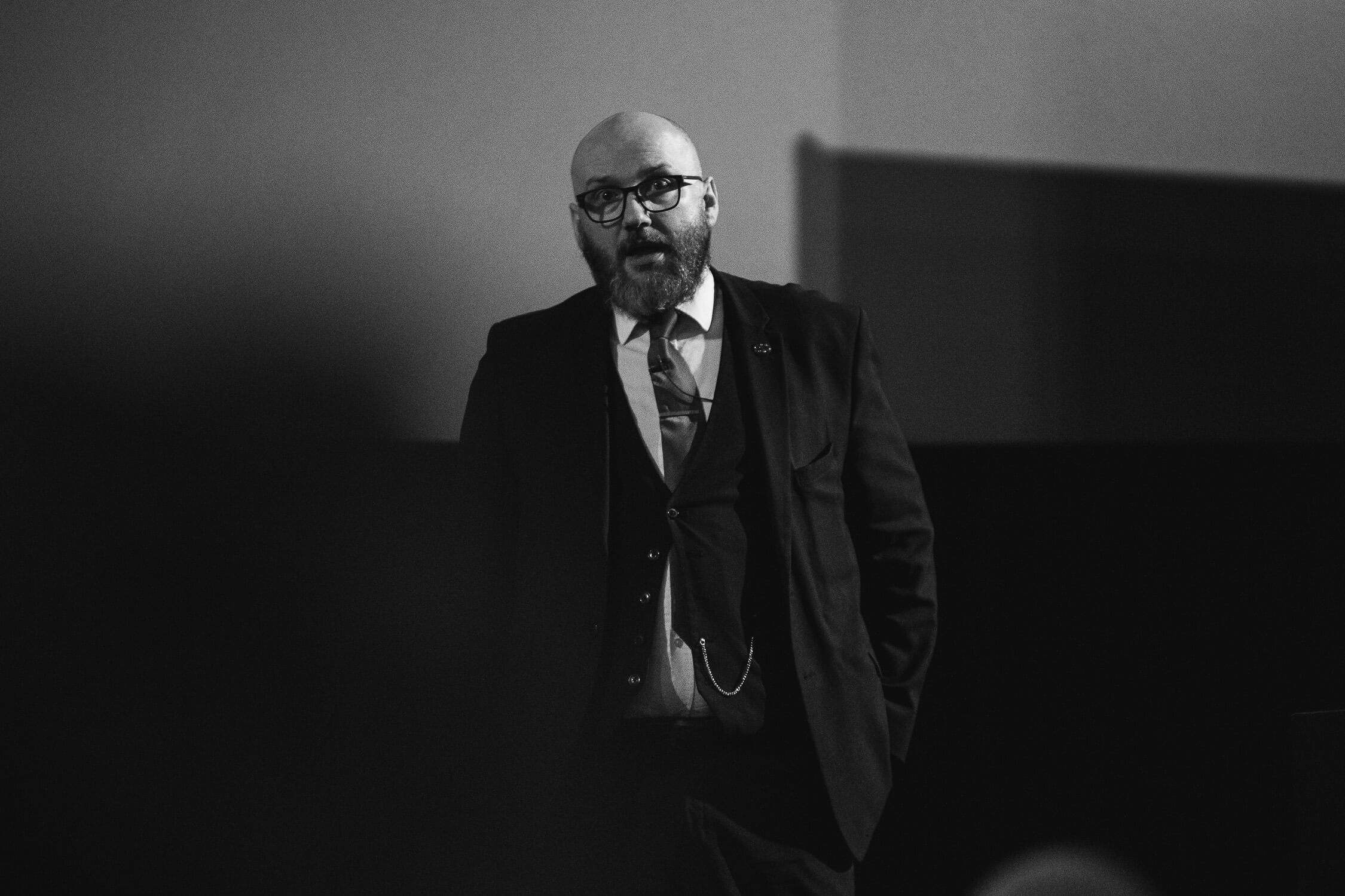 A black and white photograph of Mike Hall speaking to the crowd, dressed in a sharp suit with a pocketwatch visible